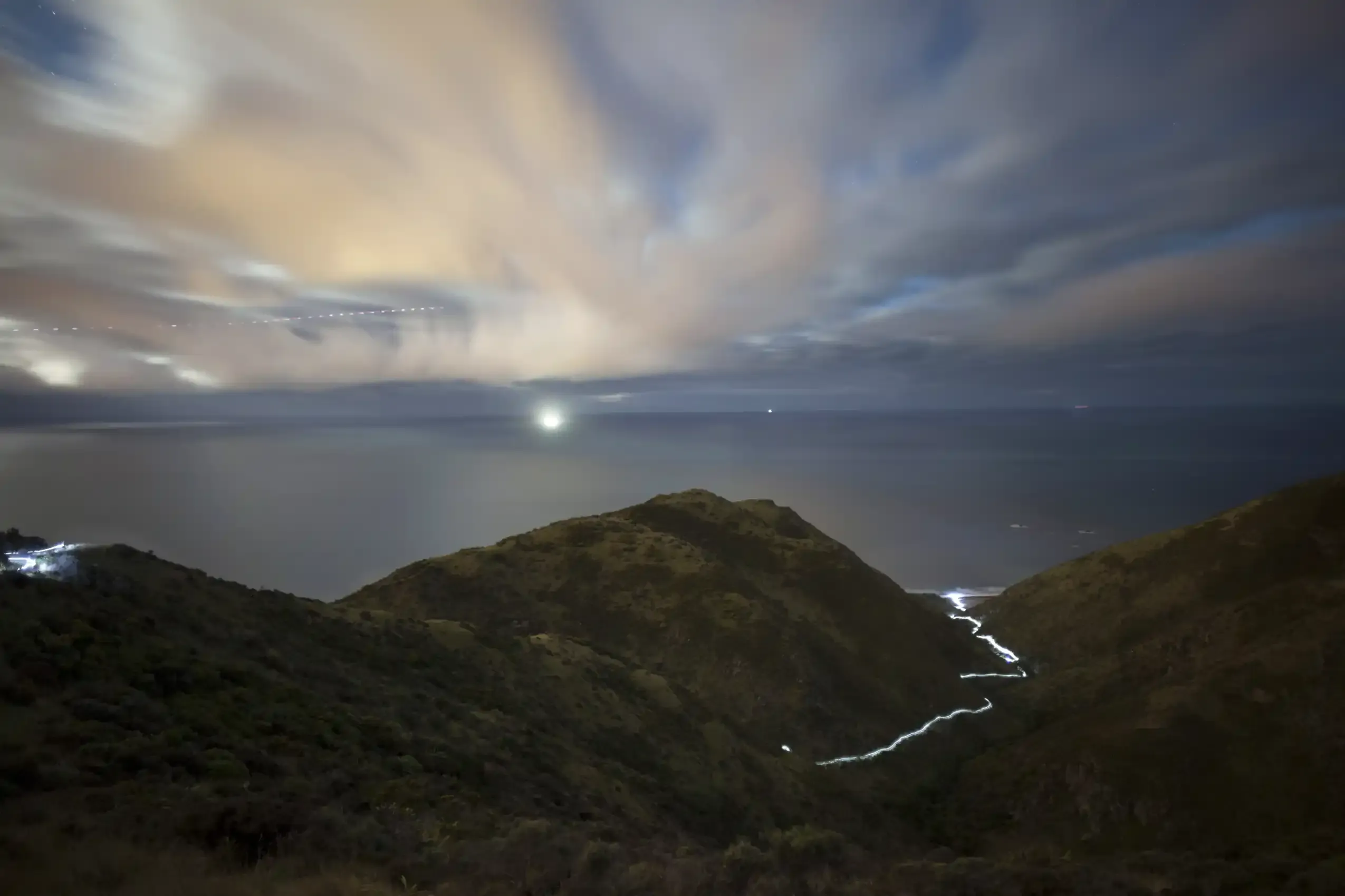 A long-exposure ribbon of light traces a coastal trail down a moonlit ridge above the sea.